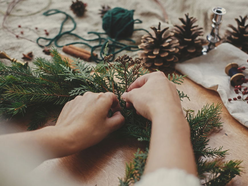 Hände mit Kräutern und Tannenzweigen, Tannenzapfen, Faden, Beeren, Zimt auf Holztisch. Weihnachtskranz-Workshop. Authentische stilvolle Stillleben