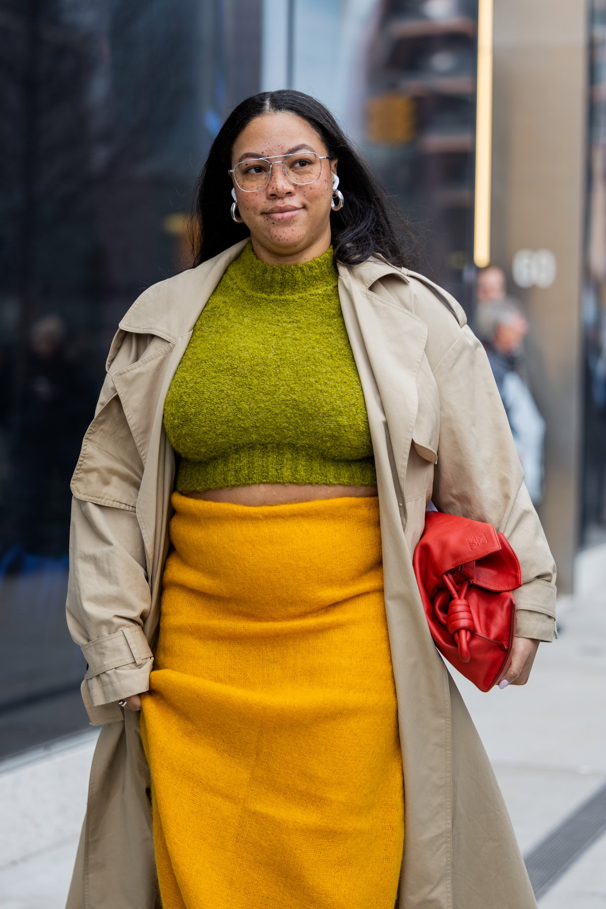 Eine curvy Frau trägt einen gelben Rock, einen Trenchcoat und einen grünen Pullover
