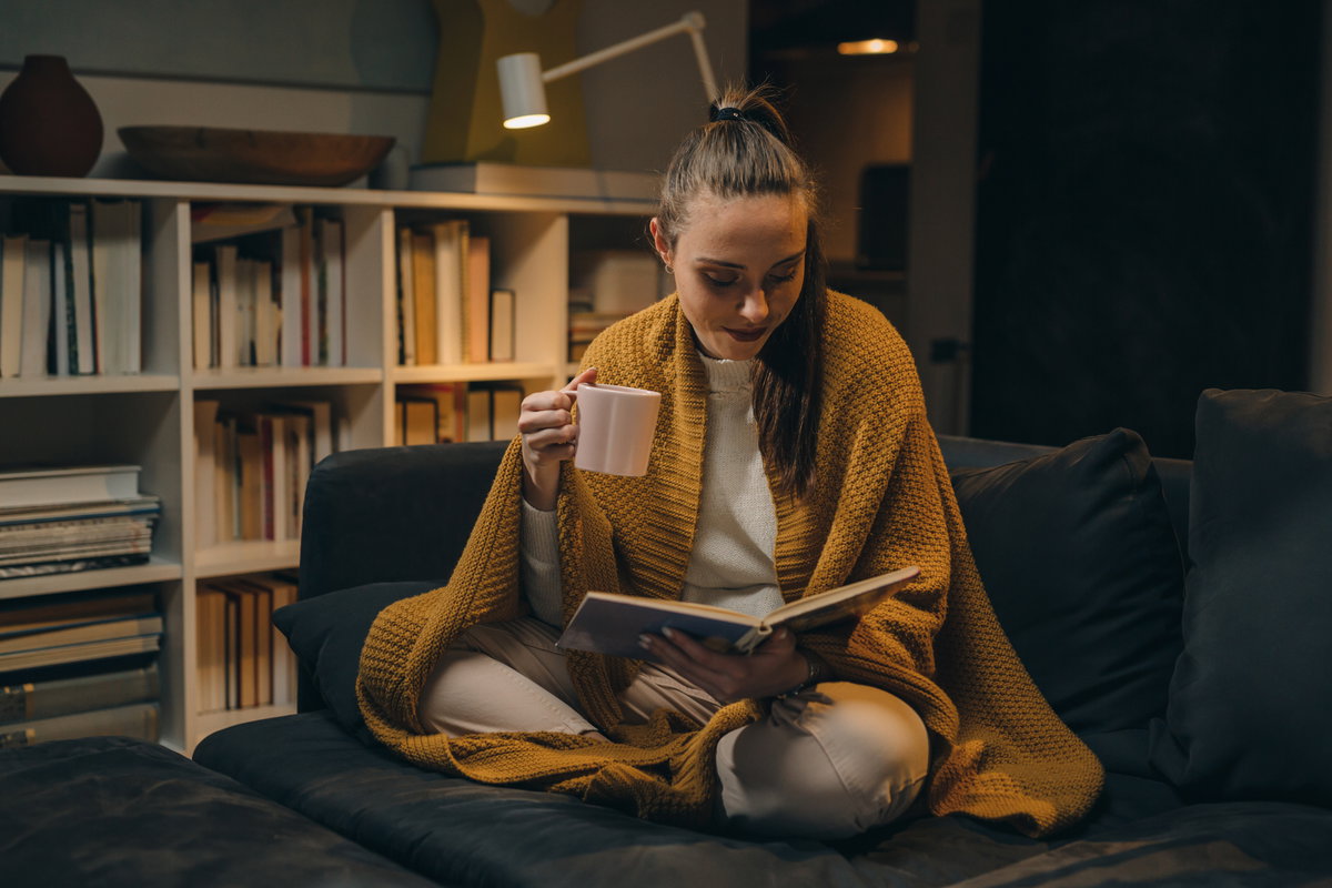 Frau mit einer gelben Decke liest ein Buch auf der Couch