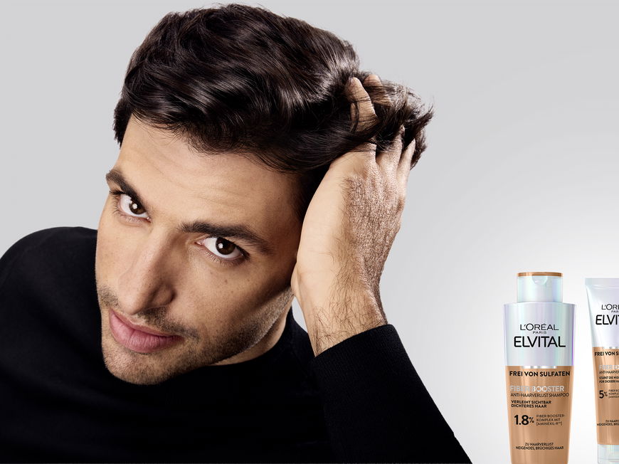Ein Mann streicht sich mit der linken Hand durch seine dunklen, kurzen Haare und neben ihm stehen Fiber Booster Anti-Haarverlust Shampoo, Spülung und Serum von L'Oréal Paris.