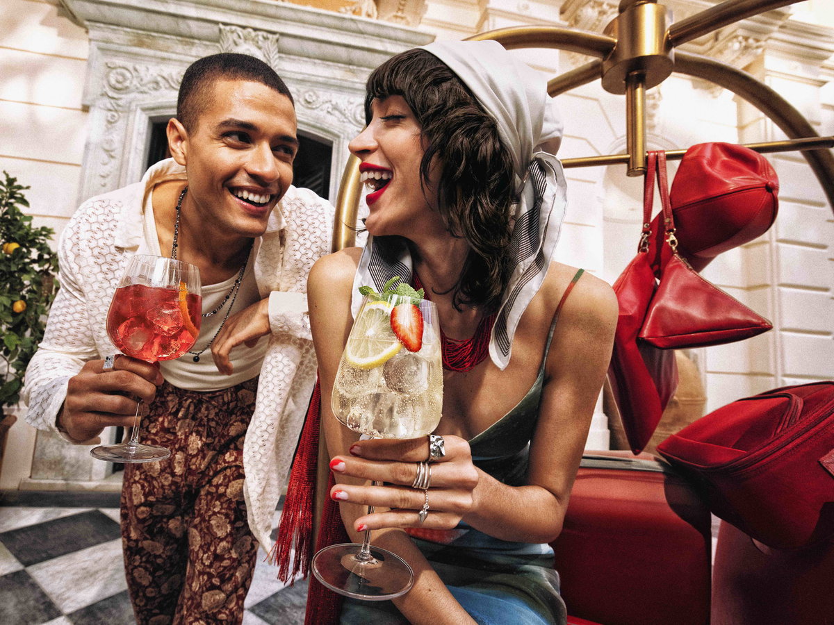 Ein junges Paar lacht gemeinsam bei einem sommerlichen Aperitivo und hält fruchtige Cocktails in einer eleganten italienischen Hotelkulisse.