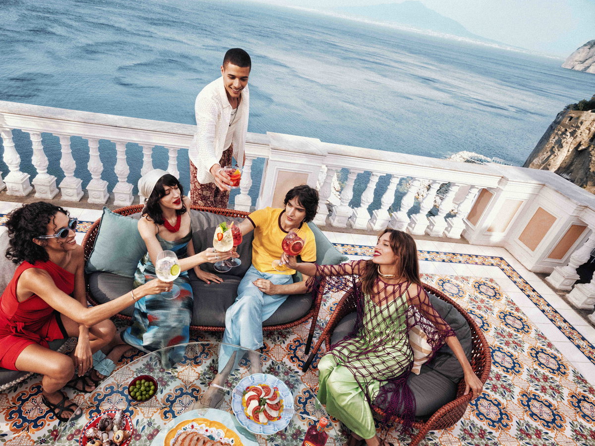 Freunde genießen gemeinsam einen italienischen Aperitivo auf einer Terrasse mit Meerblick und bunten Cocktails bei Sonnenuntergang.