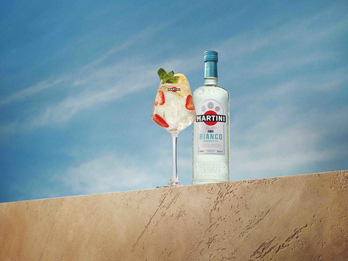 Eine Flasche Martini Bianco und ein sommerlicher Cocktail mit Erdbeeren und Minze stehen vor blauem Himmel auf einer hellen Steinmauer.