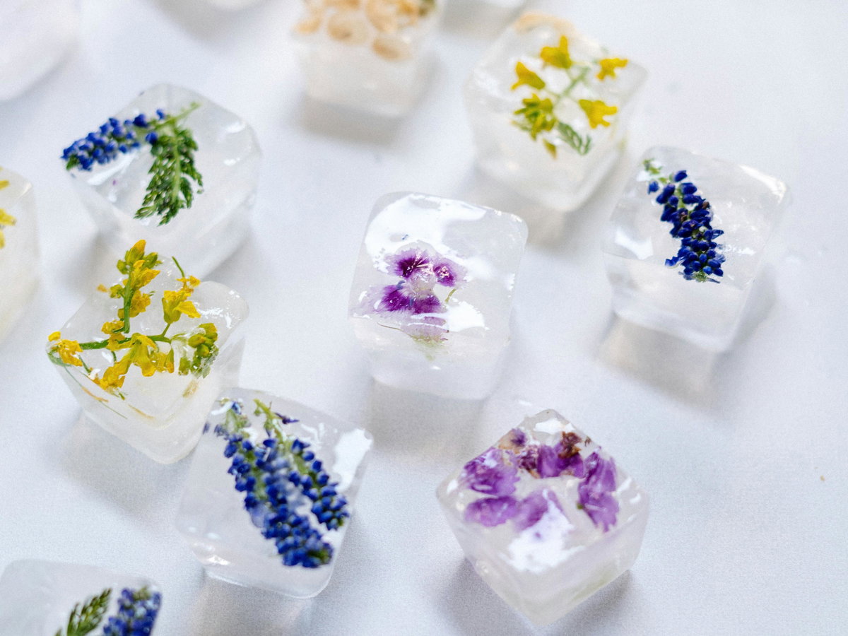 Eiswürfel mit eingefrorenen bunten Blüten liegen dekorativ auf einer hellen Oberfläche und sorgen für sommerliche Cocktailstimmung.